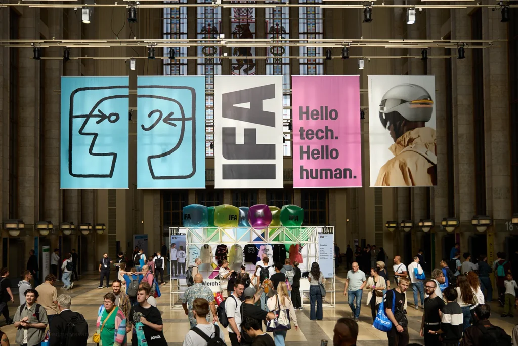 Interior hall of Messe Berlin with large hanging banners reading 'IFA' and 'Hello tech. Hello human.,' a merch stand with inflatable IFA balloons below, and crowds of visitors walking through the checkered marble floor