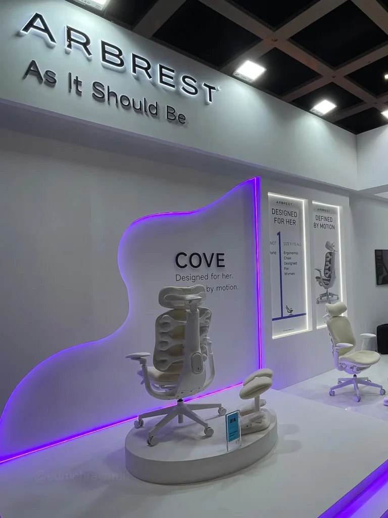 Arbrest trade fair booth with the tagline "As It Should Be" on the wall, showcasing the white "Cove" ergonomic office chair (with an exposed spine-like backrest design) on a display podium against a purple-lit curved wall panel, with additional posters reading "Designed for Her" and "Defined by Motion" and another chair displayed further back
