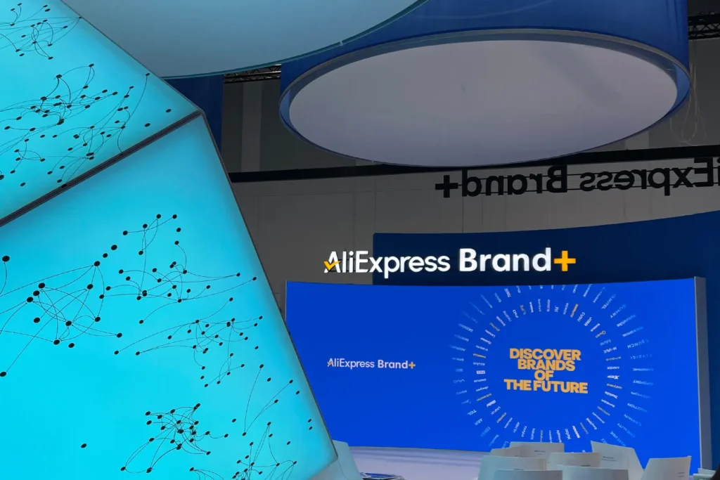 AliExpress Brand+ trade fair booth with a blue overhead sign (reading backwards from this angle), a bright cyan screen showing a network/node graphic in the foreground, and a curved blue backdrop displaying the "AliExpress Brand+" logo above a circular arrangement of brand names around the text "Discover Brands of the Future.