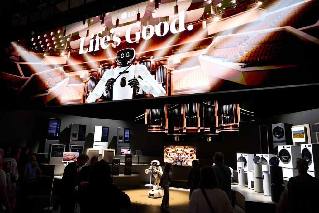 LG "Life's Good" trade fair booth at night, featuring a large overhead screen showing an animated robot conducting an orchestra, with a matching small humanoid robot on a lit stage below, surrounded by LG appliances (refrigerators, washers, speakers, TVs) on display and attendees browsing in the foreground