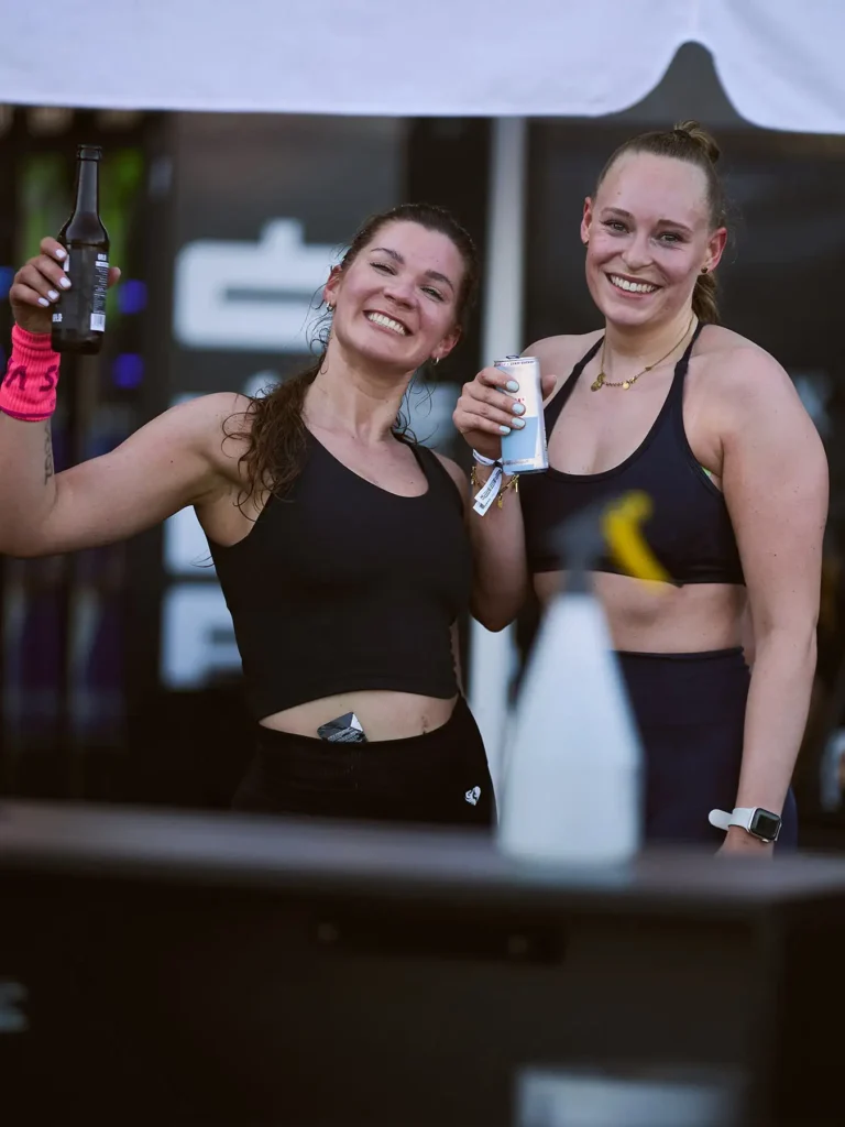 After HYROX race Magda drinking the pure and looking at the camera with her friend. she is wearing blacktop and black tight.