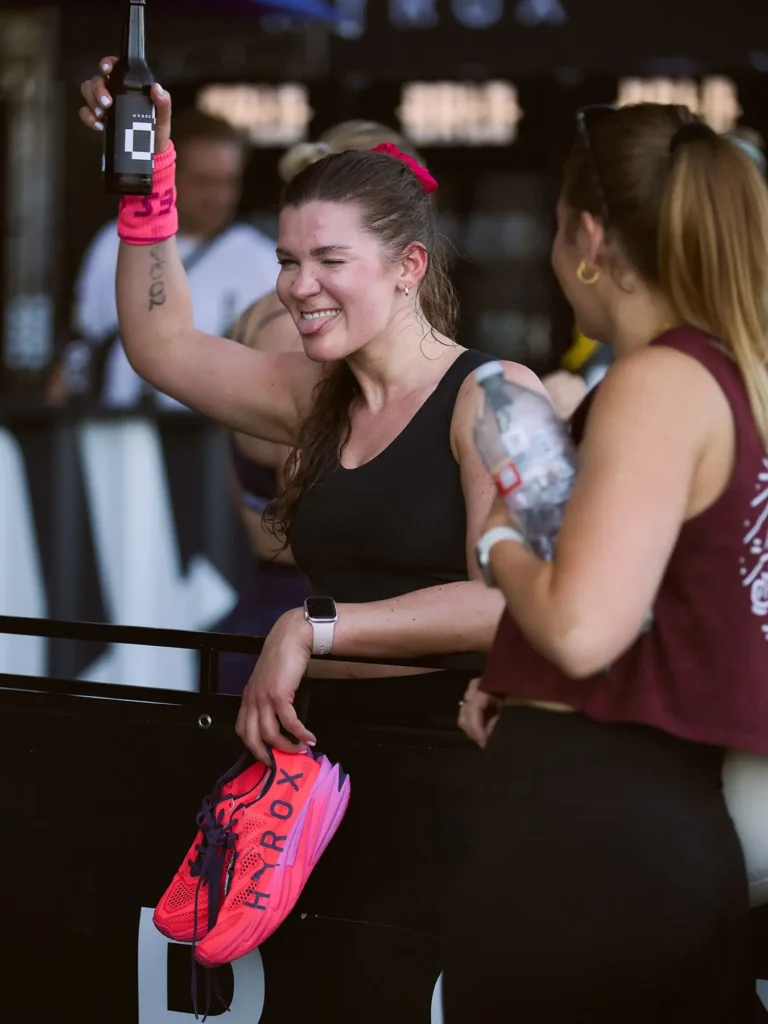 Happy after her HYROX race and looking to my camera and saying cheers and she is holding her high Rocks Puma shoes on her hand and a female friend was standing next to her