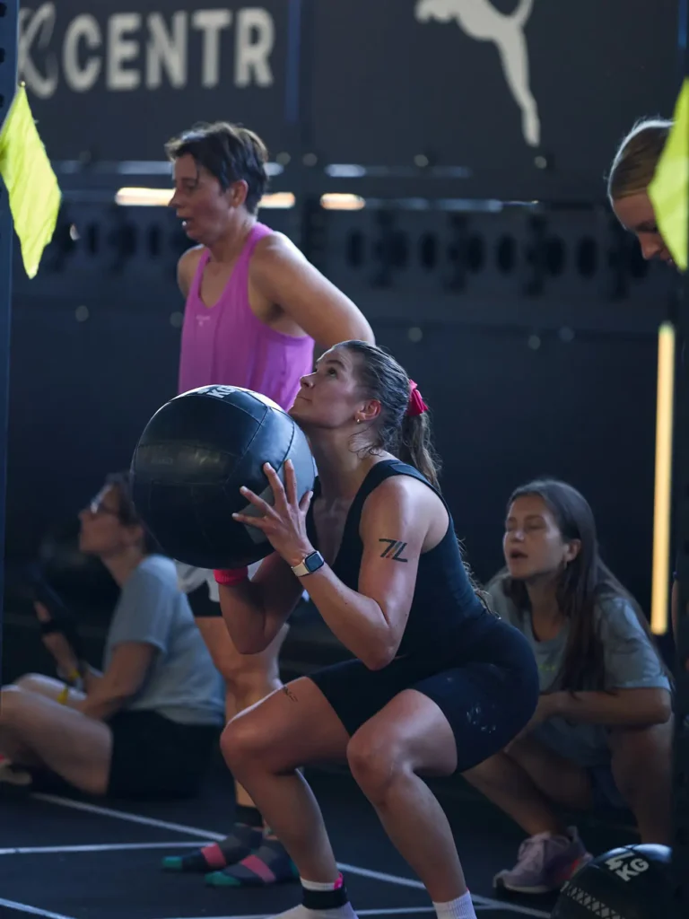 Hyrox athletes in a wall balll stations holding four kilogram ball in her hand and she is on a scout position and looking up behind her there is a judge