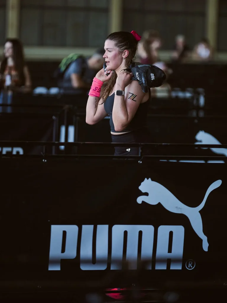 Female hyrox athlete holding sandbag lounge on her shoulder and in foreground you can see the white Puma logo