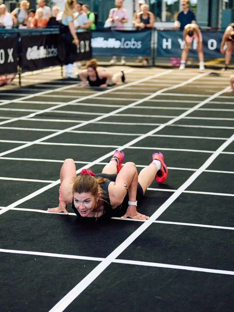 Burpee broad jump station in HYROX Berlin Tempelhof. a female touching ground with chest and ready to stand up the athlete called Magda, she is wearing red Puma HYROX shoes and watching front