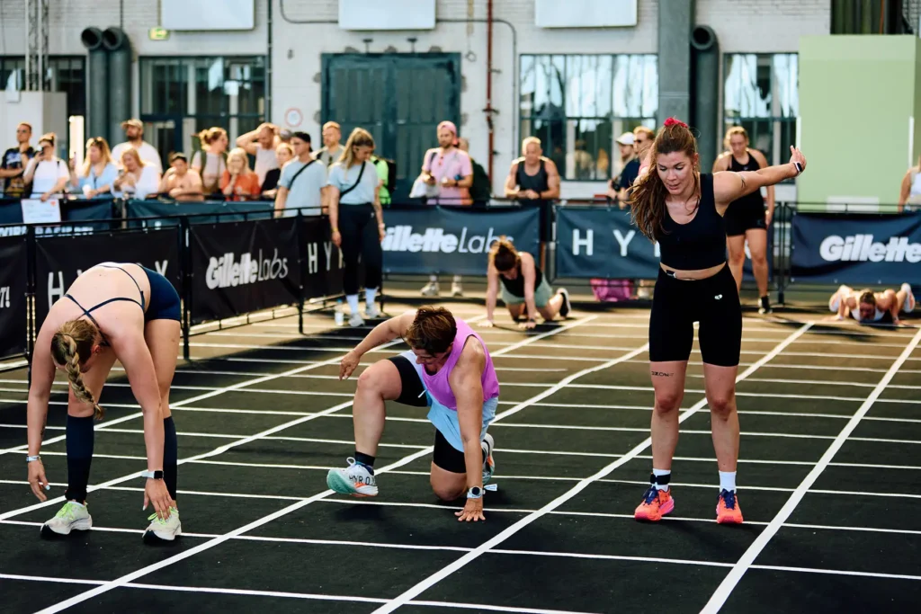 Burpee broad jump station in HYROX Berlin Tempelhof. there is three female athletes in front and there is three female atlas in background and two judges are watching them and people are watching them
