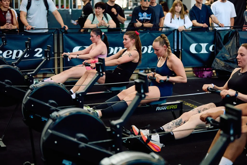 Rowing station in HYROX race. In the picture there is four HYROX female athletes visible and there are ruling and the people watching them