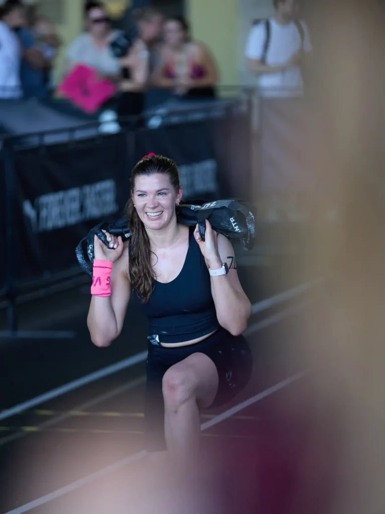 This is sandbag lunge station in HYROX race. Magda careing a 10 kg sandbag lunge on her shoulders at HYROX Berlin. She's looking to the camera and smiling