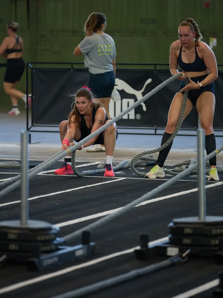 Magda crouching low to pull a weighted sled with a rope at HYROX Berlin 2026, alongside another athlete in the adjacent lane.